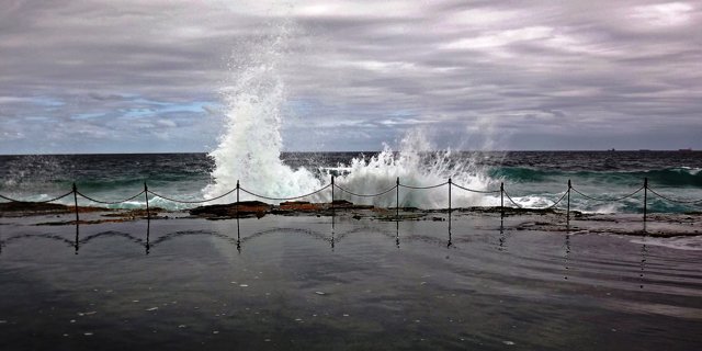 A breaking wave rises beyond chain-linked posts at the edge of a rock pool.