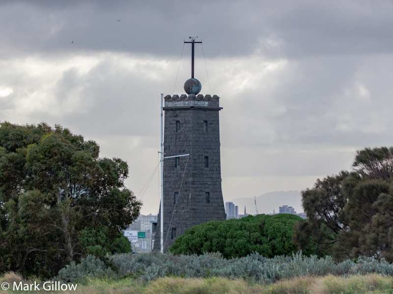 The stone timeball tower rises among shrubs, with a distant city skyline behind it and a cloudy sky.