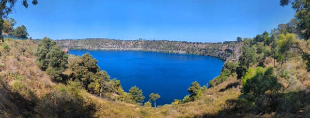 Blue water enclosed by steep vegetated crater slopes beneath a clear sky, seen in a broad panoramic view.