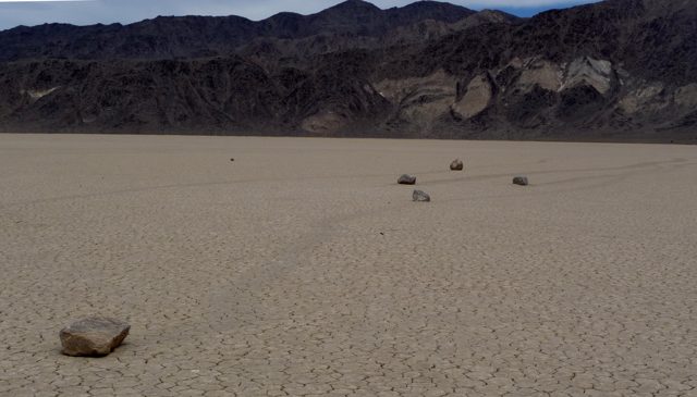 Stones and faint trailing furrows cross a flat cracked lakebed below dark desert mountains.
