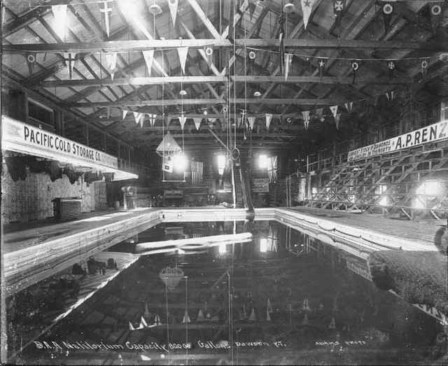 Black-and-white photograph of an indoor swimming pool beneath timber roof trusses, with hanging flags and stepped seating; the photographer’s inscription remains along the bottom.
