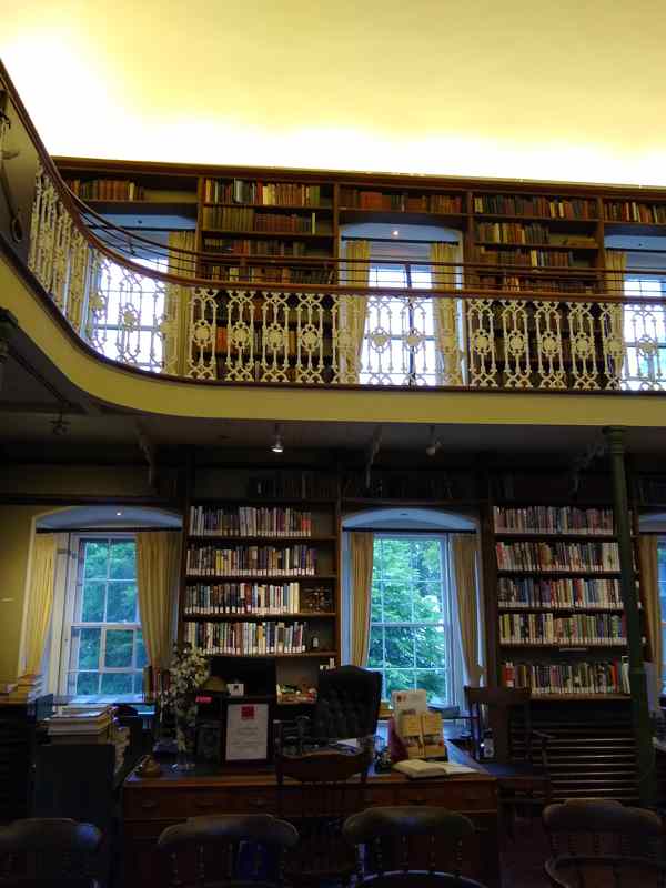 Bookshelves line two levels of a library, with an ornate pale gallery railing, windows and a desk below.