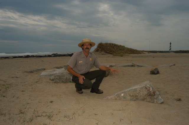 A ranger crouches beside partly sand-covered foundation blocks; the sea lies to the left and the relocated lighthouse stands in the distance.