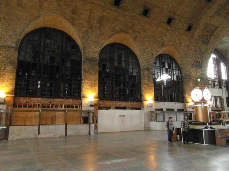 A tall arched terminal interior, with an illuminated freestanding clock at right and a few visitors beside it.