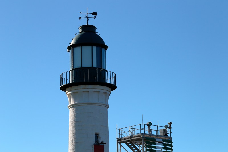The upper white lighthouse, dark lantern and weather vane stand against blue sky, beside a steel platform with lights and equipment.