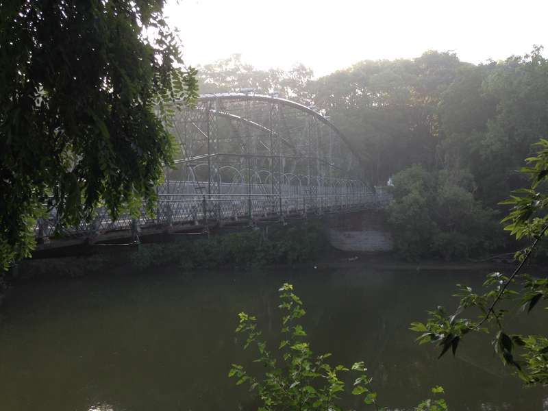 The arched bridge and lattice railings seen through riverside trees in hazy morning light.