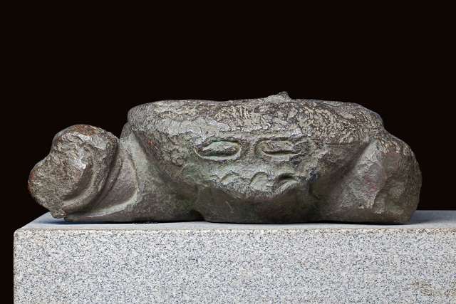 A low greenish bronze crab fragment with worn shell and truncated limbs, displayed on a pale stone plinth against a dark background.