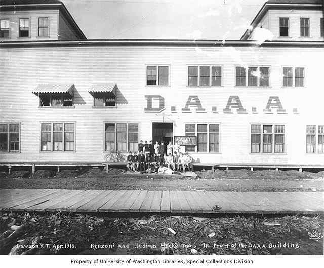 A black-and-white group photograph of hockey players outside a large wooden building marked D.A.A.A., with a boardwalk in front, a photographer inscription and a collection credit strip below.