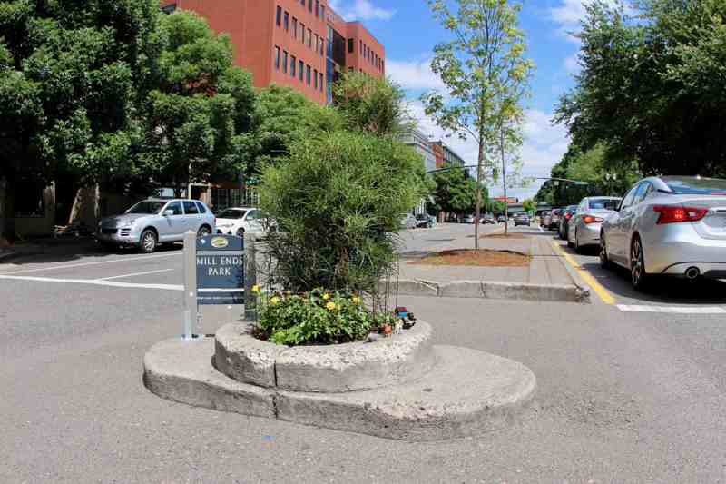 A small circular planting with yellow flowers, miniature objects and a Mill Ends Park sign stands between traffic lanes.