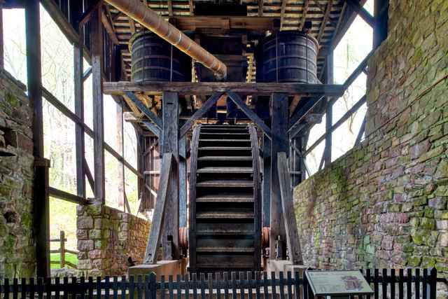 A large wooden waterwheel beneath two barrel-shaped blowing tubs inside a stone-and-timber wheelhouse.