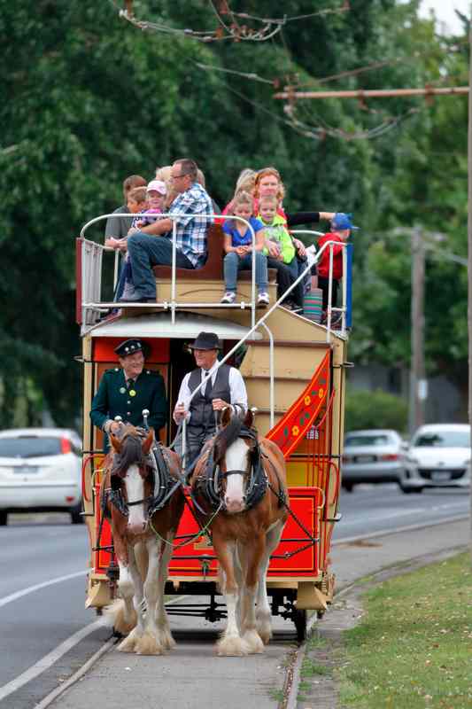 Two horses harnessed to a red-and-cream double-deck tram, with passengers in the open upper seating area beside a road.