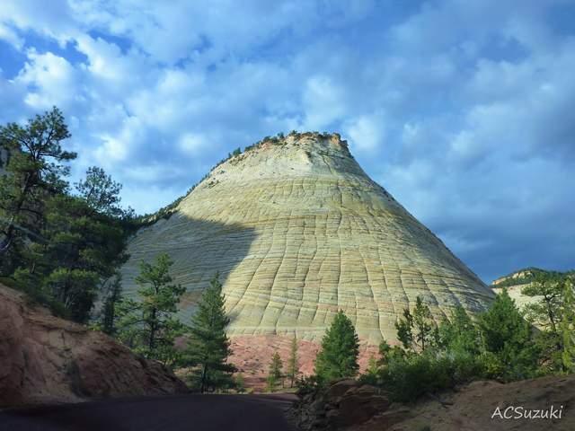 A pale sandstone mountain with crossing surface lines rises above trees and a road under a cloudy sky; the photographer’s ACSuzuki credit remains at lower right.