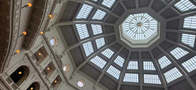 An upward, angled view of pale dome ribs, glazed roof panels and an octagonal central lantern, with gallery lights at left.
