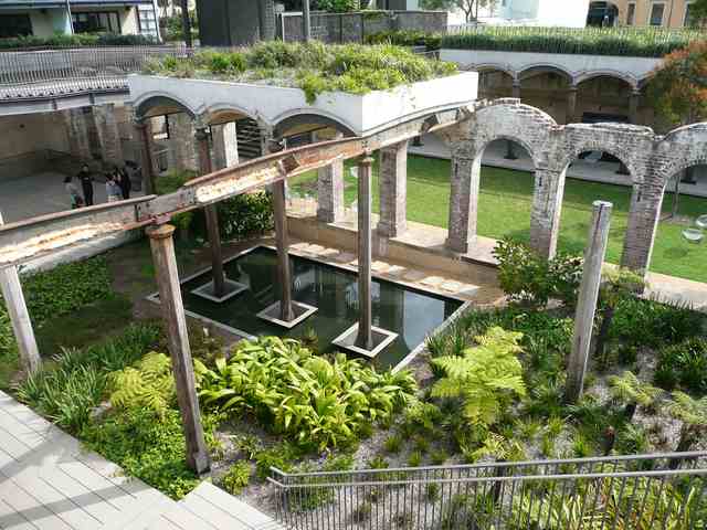 A pond reflects vertical supports beneath fragments of brick arches, beside newer curved concrete edges, planted beds and a walkway.