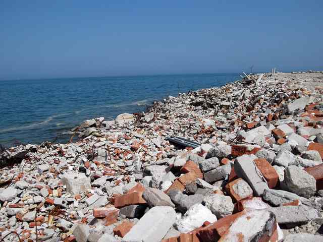 Broken concrete, red bricks and metal reinforcement cover a shoreline beside open lake water.