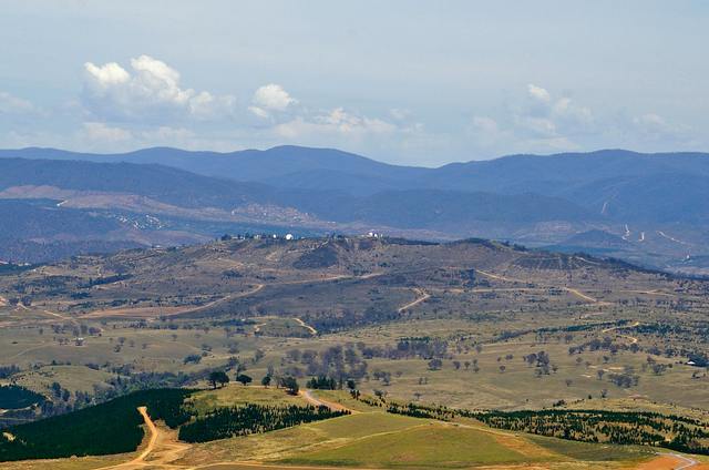 A low ridge with small white observatory domes above fields and tree plantations, with higher ranges behind it.