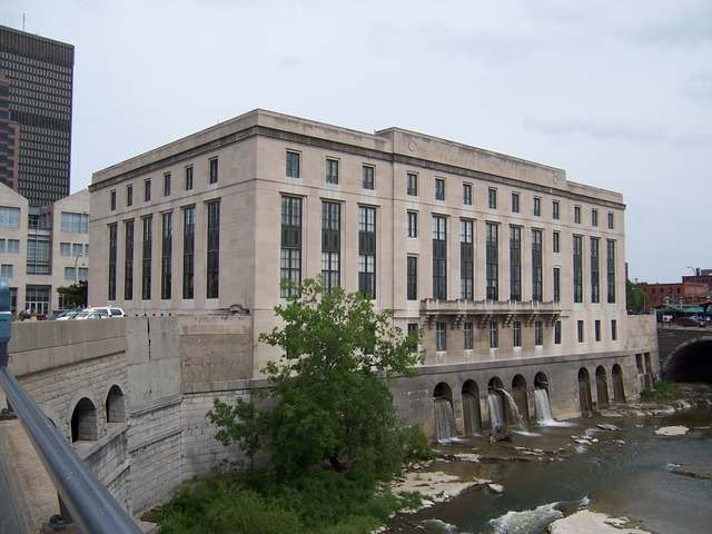 A pale stone library above a river, with tall rectangular windows and water spilling from arches below the building.