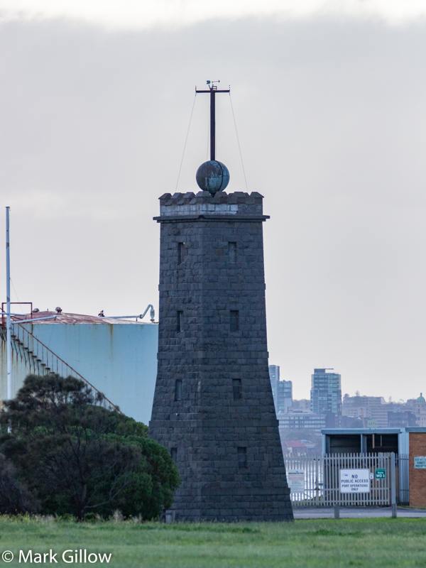 A square grey stone tower with battlements and a ball on a tall mast, beside port buildings and a fence.