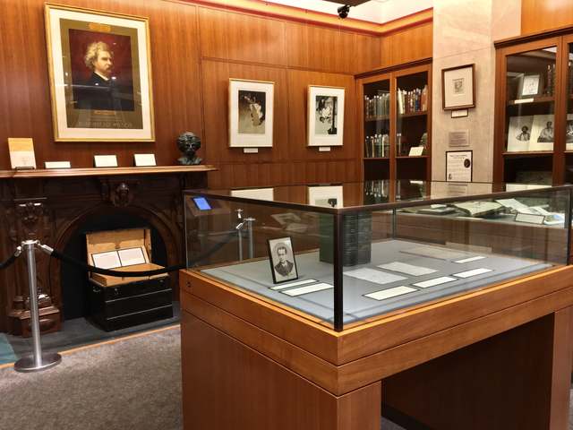Wood-panelled exhibition room with glass display cases, books, framed images and a fireplace.