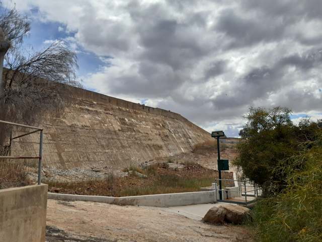 A large sloping dam wall stands behind a path, railings and small signs, with trees to the right beneath a cloudy sky.
