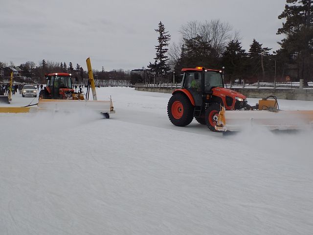 Two red tractors with broad front attachments clear snow from the canal; small figures and another vehicle are farther behind.