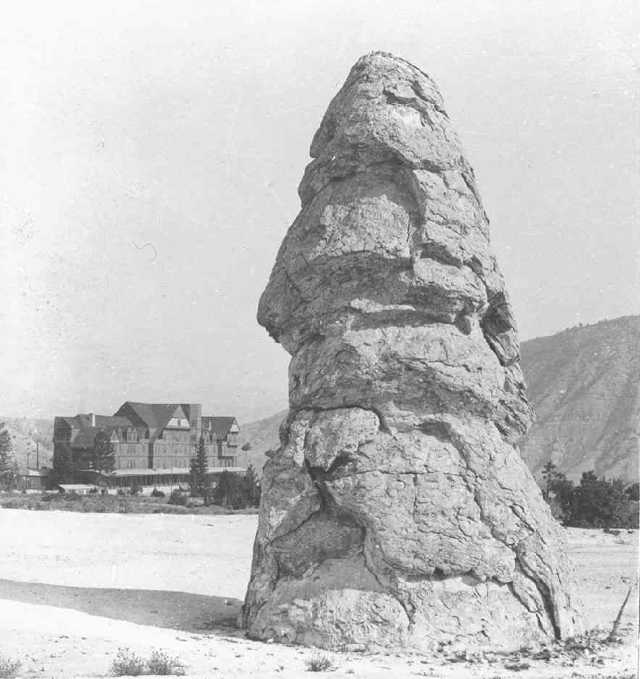 An upright layered rock cone stands in front of a large hotel in a black-and-white photograph.