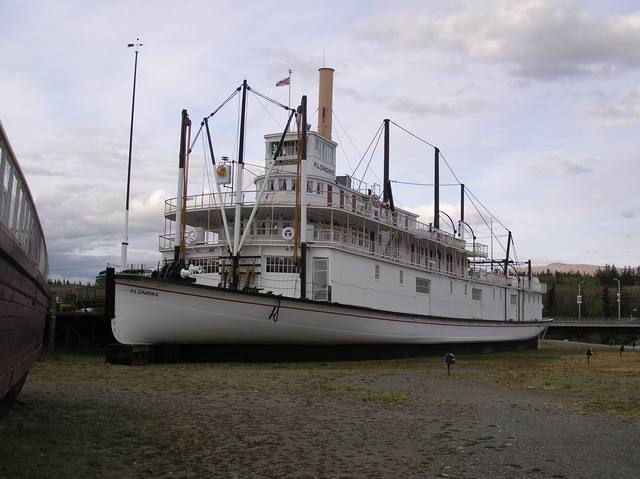 A white sternwheeler rests on land, with its bow, decks, wheelhouse, funnel and rigging visible.