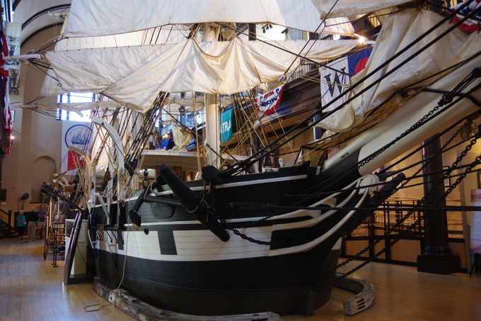 The black-and-white bow of a ship model, crossed by chains and rigging, stands indoors beneath white sails; small visitors are visible far to the left.