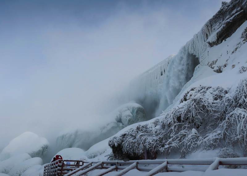 Ice coats the cliff and vegetation beside falling water; a snow-covered viewing rail crosses the foreground.