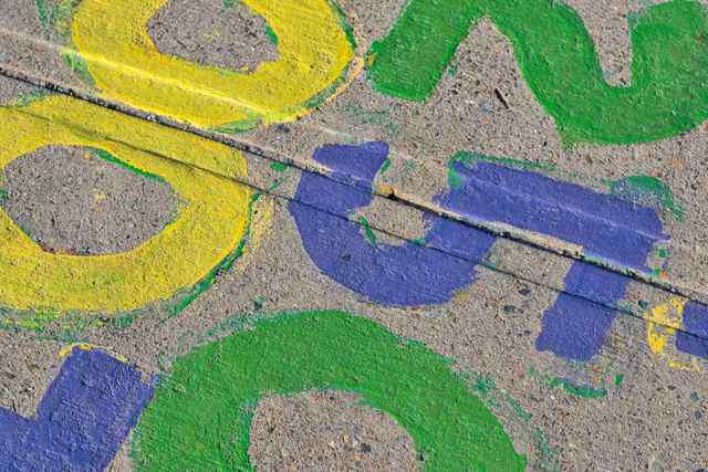 Yellow, green and purple painted shapes overlap on rough concrete, crossed by a diagonal pavement joint.