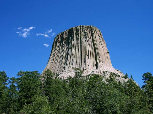 The columned rock formation rises above a green wooded foreground beneath blue sky.