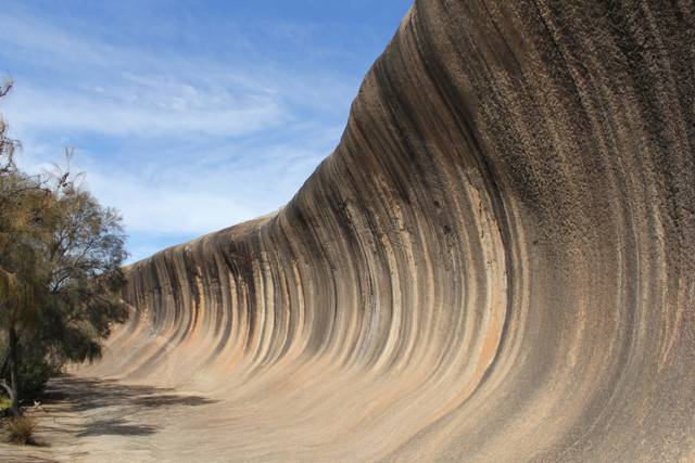 The granite face curves outward above bare ground, with dark and tan vertical streaks and trees at the left.