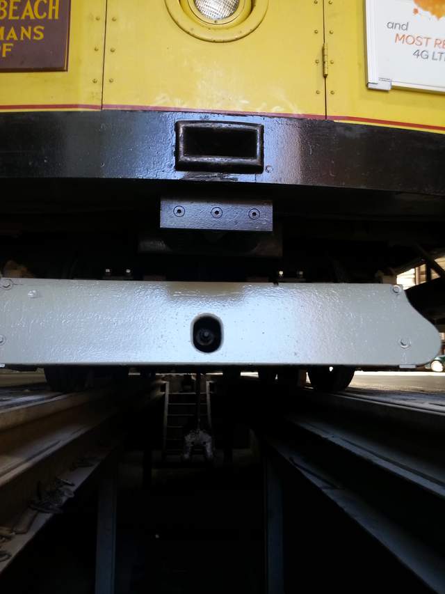 The yellow front of a cable car above a dark maintenance pit, with part of its grip extending below the undercarriage.