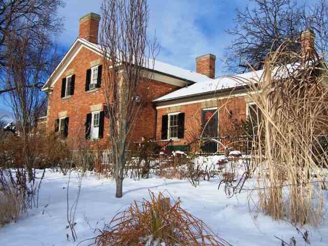 A red-brick house with dark shutters and several chimneys stands behind a snow-covered garden and leafless trees.