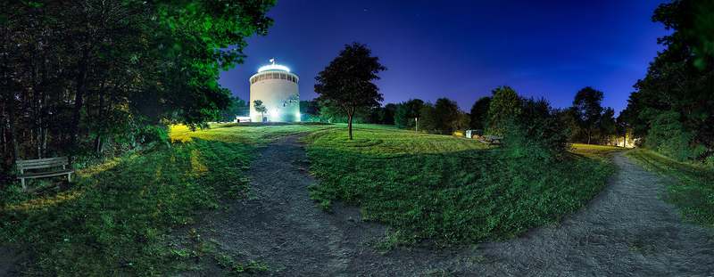 An illuminated cylindrical standpipe beyond grass, trees and paths in a wide night view.
