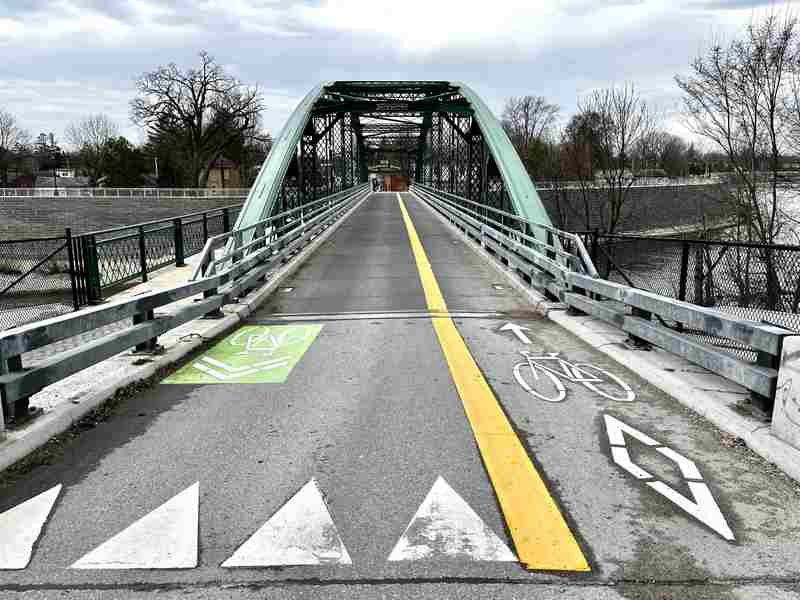 A green arched iron bridge seen along its roadway, with bicycle markings, a yellow line and a separate walkway at the left.