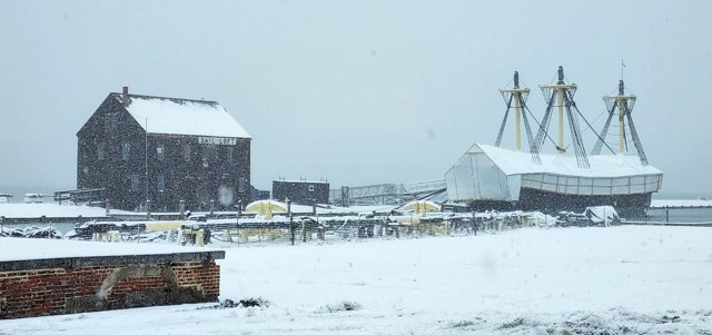 A white cover encloses the ship at a snowy wharf, with three lower mast sections above it and a dark Sail Loft building to the left.
