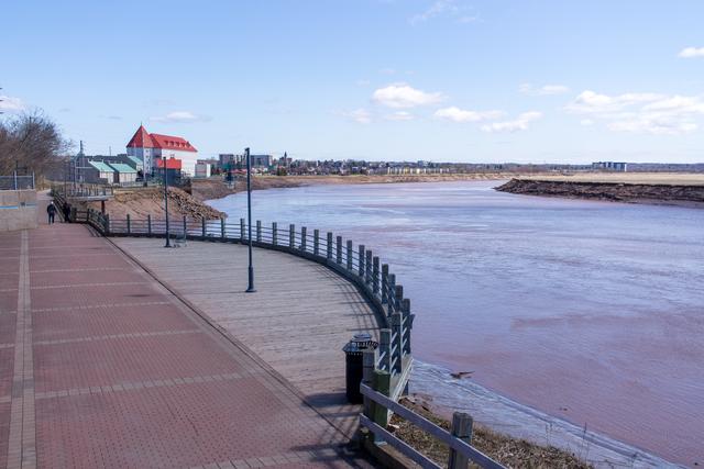A curving waterfront walkway and railings beside the Petitcodiac River, with buildings and two distant pedestrians.