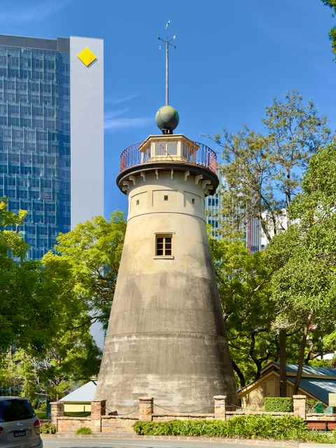 A cylindrical stone tower with a balcony and tall roof-mounted pole stands among trees and modern city buildings.