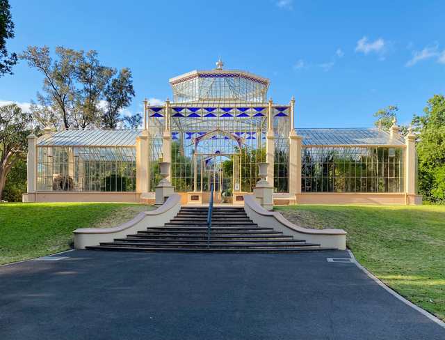 A symmetrical glasshouse with pale framing and blue geometric panes stands above broad steps and grassy slopes beneath a blue sky.