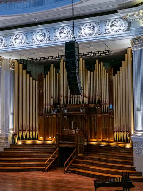Golden-coloured organ pipes and dark wooden casework rise behind a suspended black speaker.