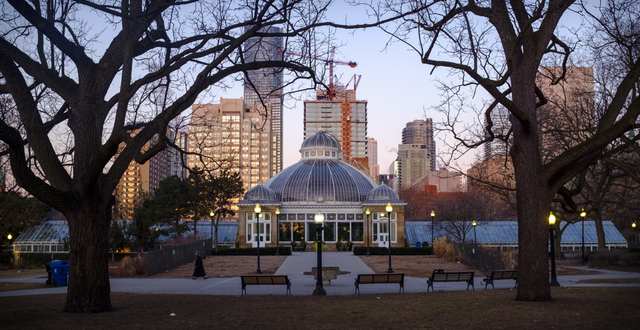 A greenhouse with three domes and a central entrance, framed by bare trees with tall city buildings behind.