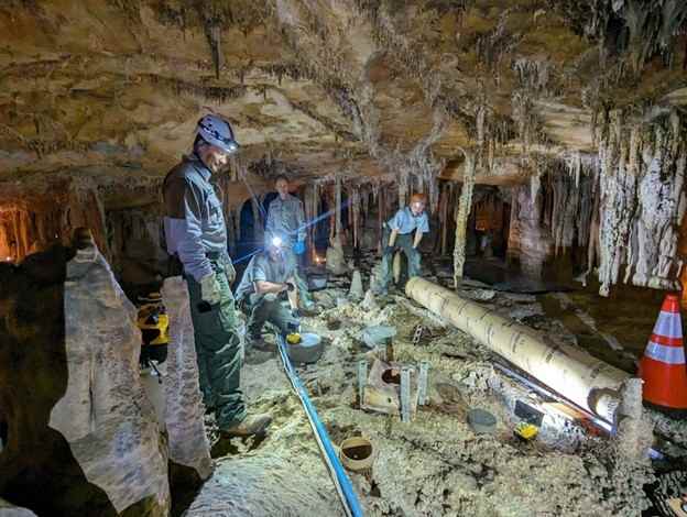 Helmeted workers among exposed cave-floor material and construction equipment beneath hanging cave formations.
