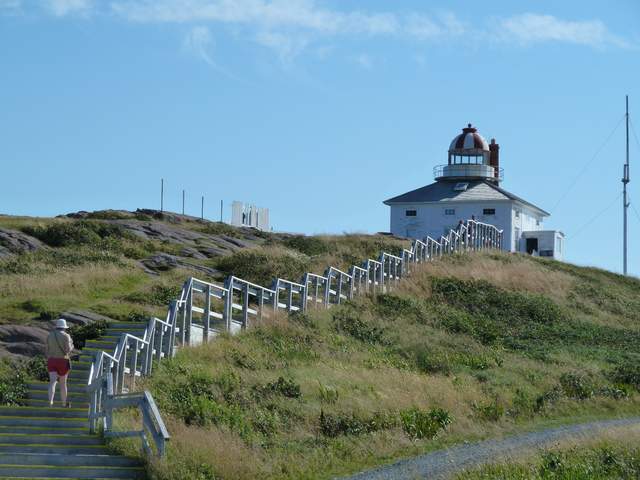 A square white lighthouse with a central domed lantern stands above a hillside stairway with small visitors.