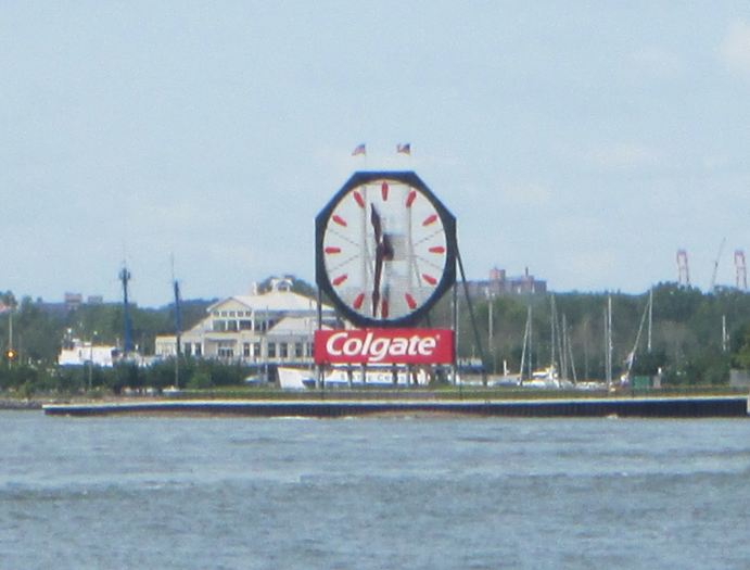 Across a river, a large octagonal clock with a red Colgate sign stands in front of marina buildings and boat masts.