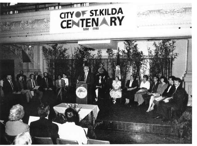 A black-and-white photograph shows a speaker and seated guests on a stage beneath a city-centenary banner, with an audience in front and decorated balcony above.