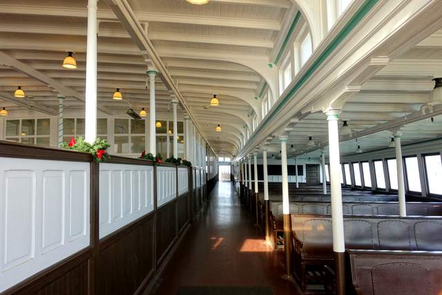 An empty ferry interior with rows of wooden benches, slender columns and a long passage beside windows.