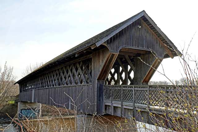 Oblique view of the roofed wooden bridge, its lattice side, entrance and river below.
