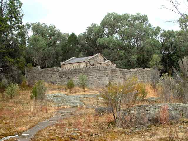 A small stone building rises behind a high enclosing stone wall, with trees and exposed rock around it.