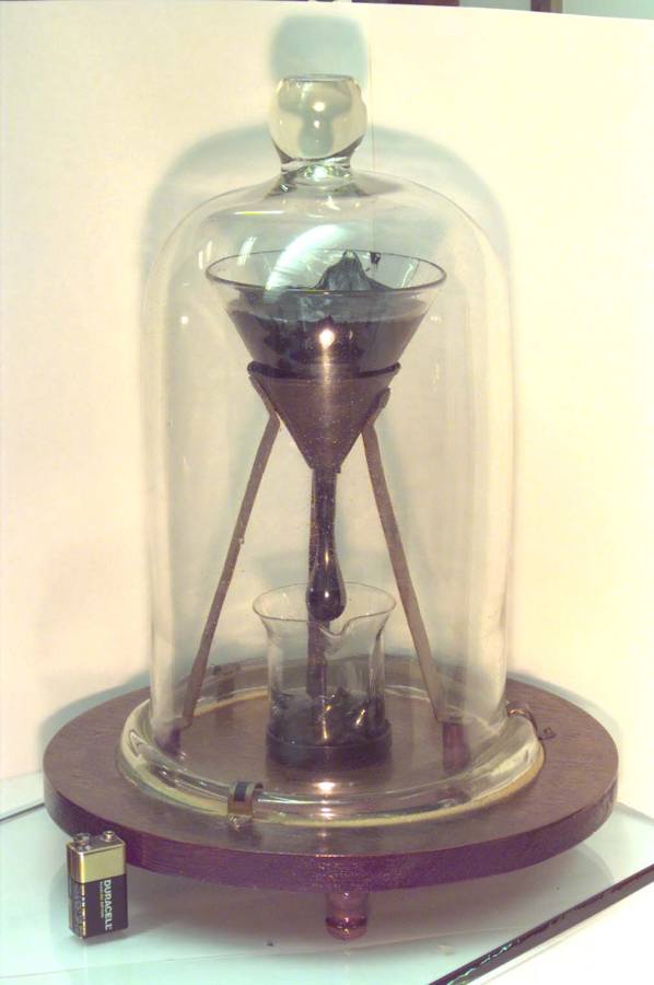 A funnel and hanging pitch stand above a collecting beaker inside a bell jar; a battery sits beside the wooden base.